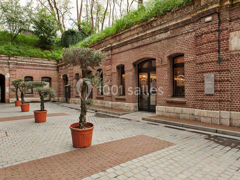 Cour pavée bordée de bâtiments en briques rouges avec des arbres en pots et une végétation sur les toits.