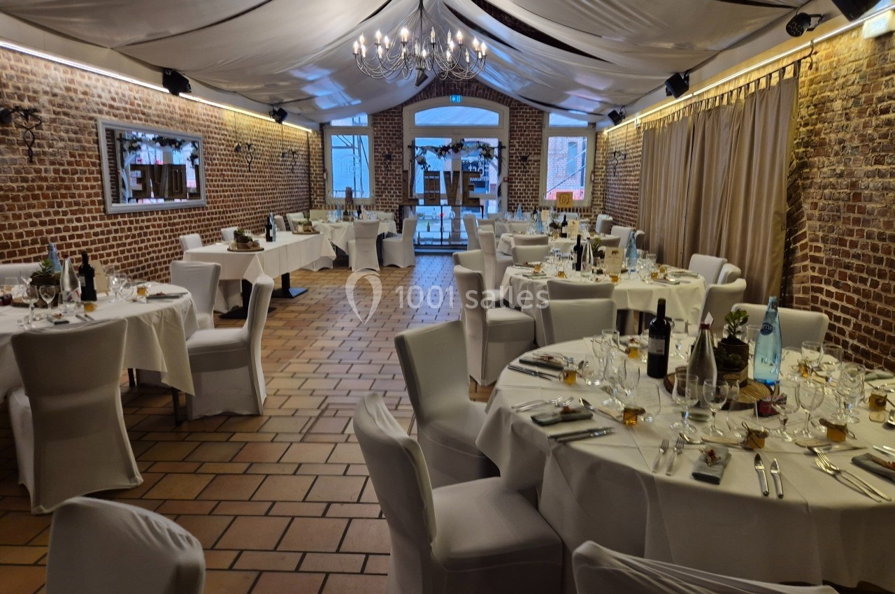 Salle de réception décorée avec des tables rondes dressées, nappes blanches et lumière tamisée sous un plafond voilé.