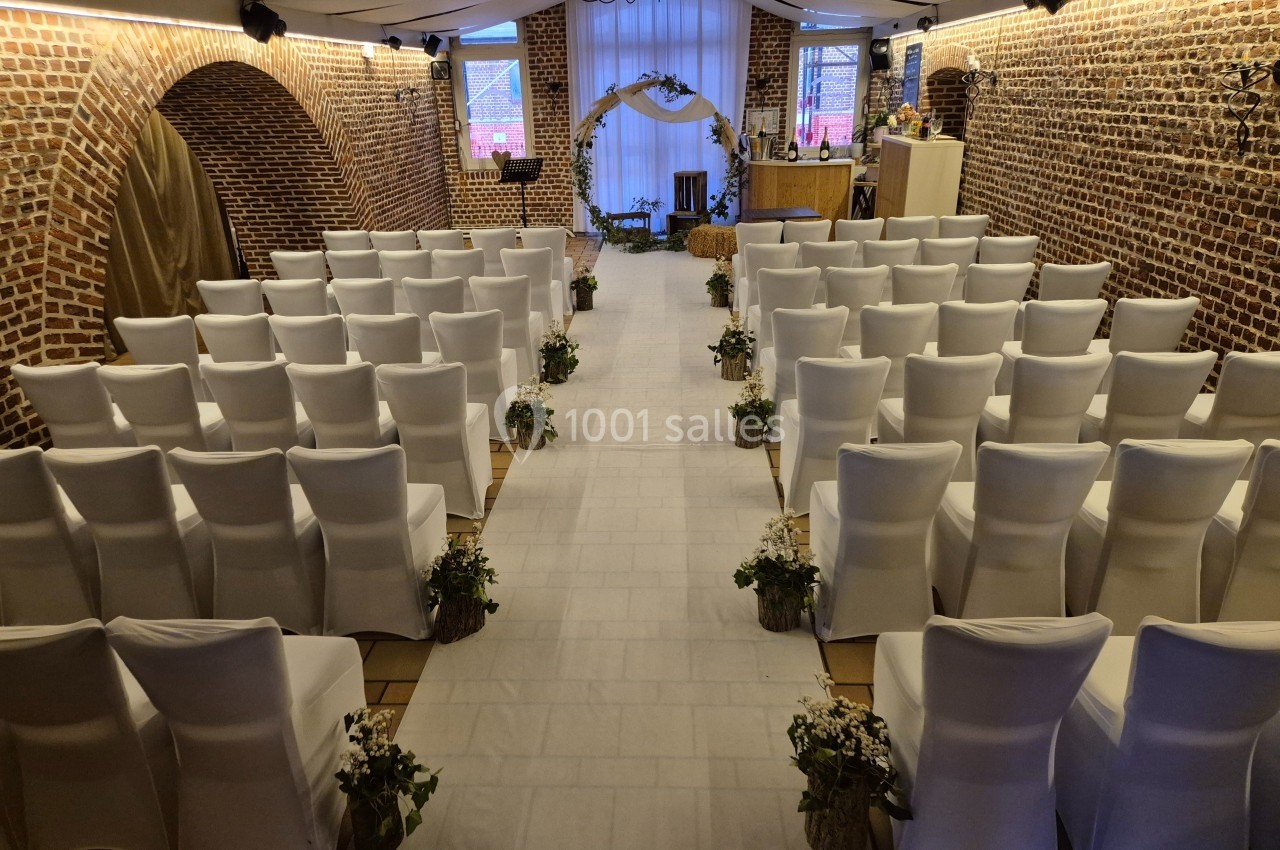 Salle de cérémonie décorée avec des chaises blanches alignées, une arche fleurie et des murs en briques apparentes.