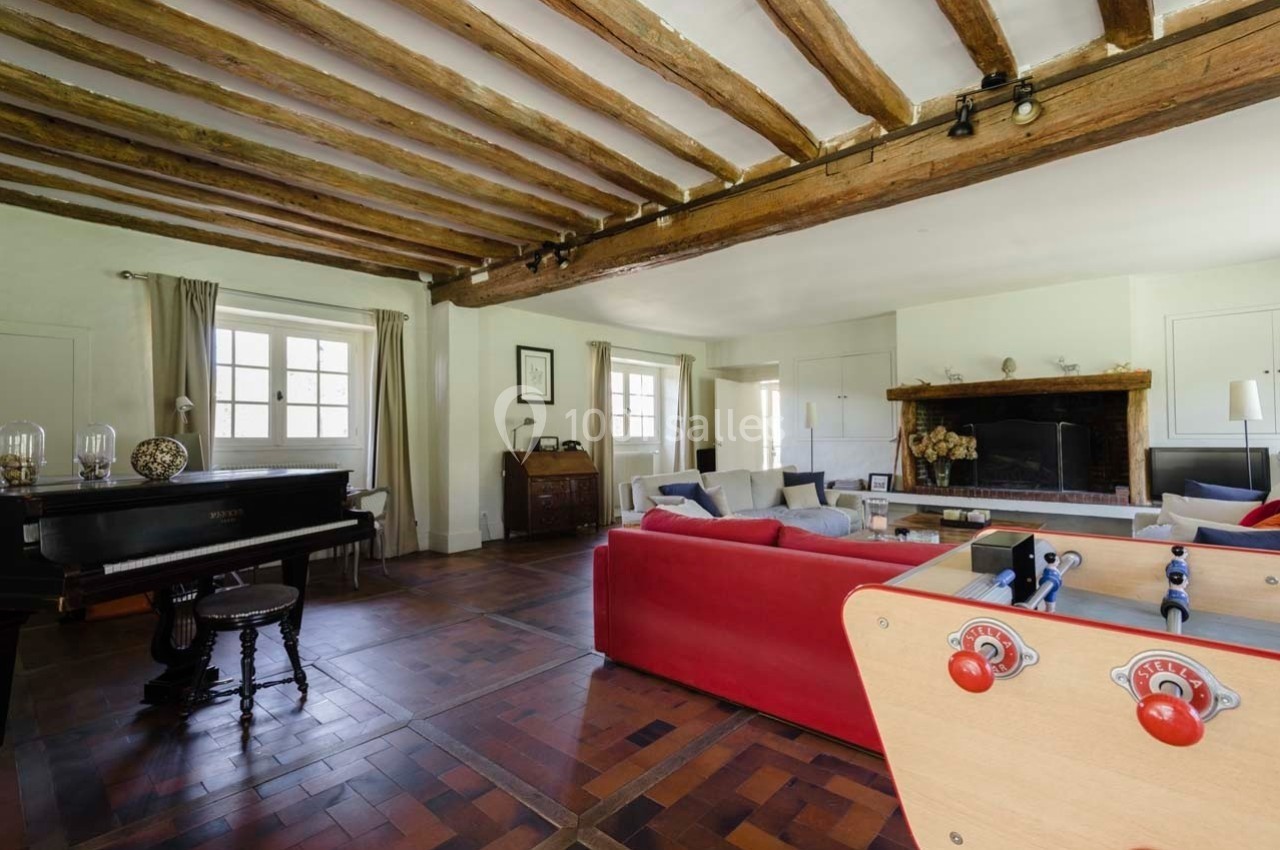 Salon spacieux avec poutres apparentes, cheminée, canapé rouge, piano à queue et baby-foot sur un sol en parquet.