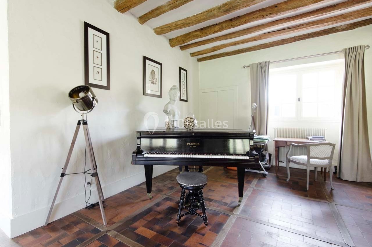 Salle lumineuse avec un piano noir, tabouret, trépied vintage, bureau et fenêtres encadrées de rideaux beiges.