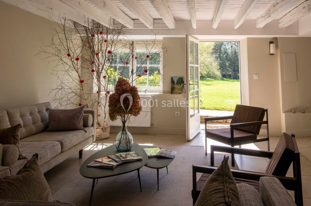 Salon lumineux avec poutres apparentes, mobilier moderne, grande baie vitrée ouverte sur un jardin verdoyant.