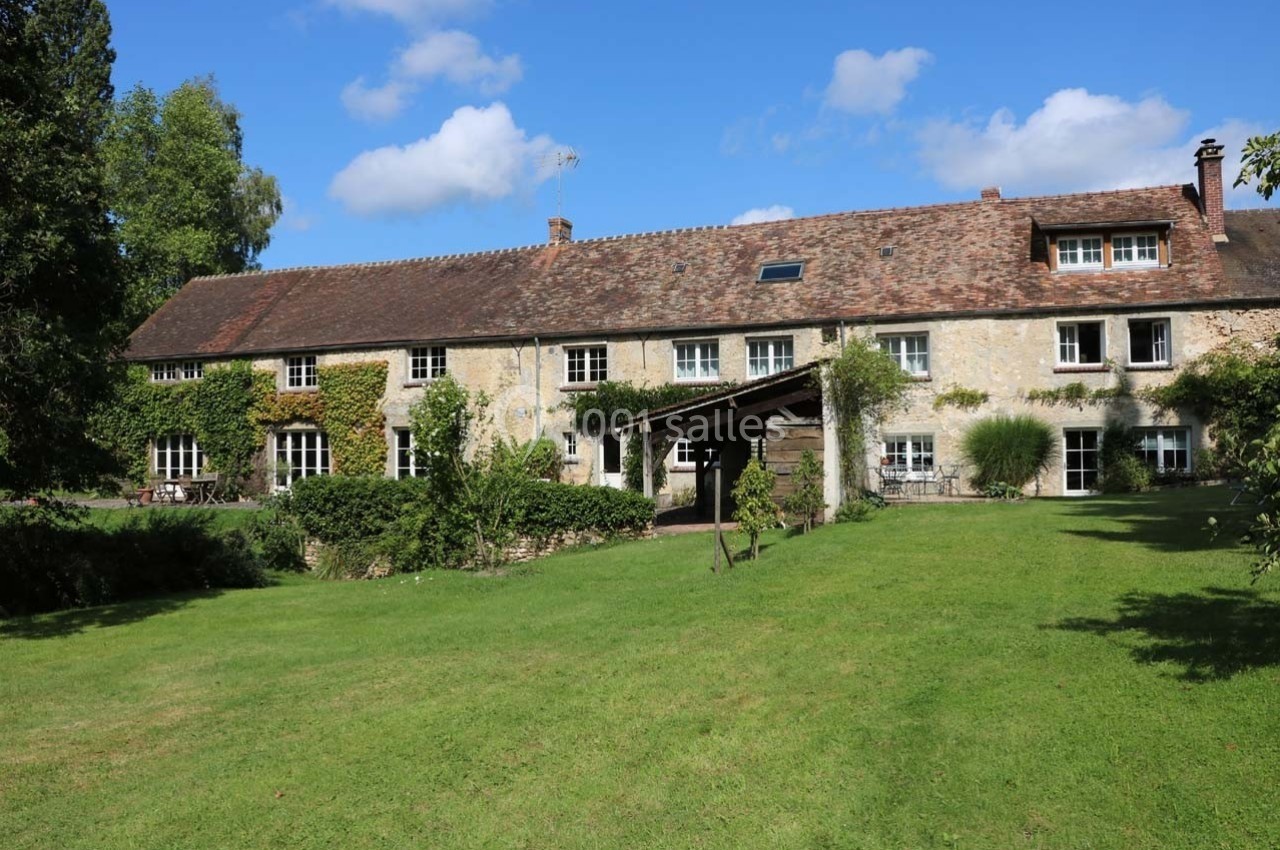 Grande maison en pierre avec toit en tuiles, entourée de verdure et d'un jardin sous un ciel bleu.