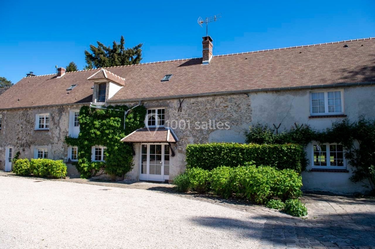 Façade d'une maison en pierre avec toit en tuiles, fenêtres blanches et végétation grimpante, sous un ciel bleu.