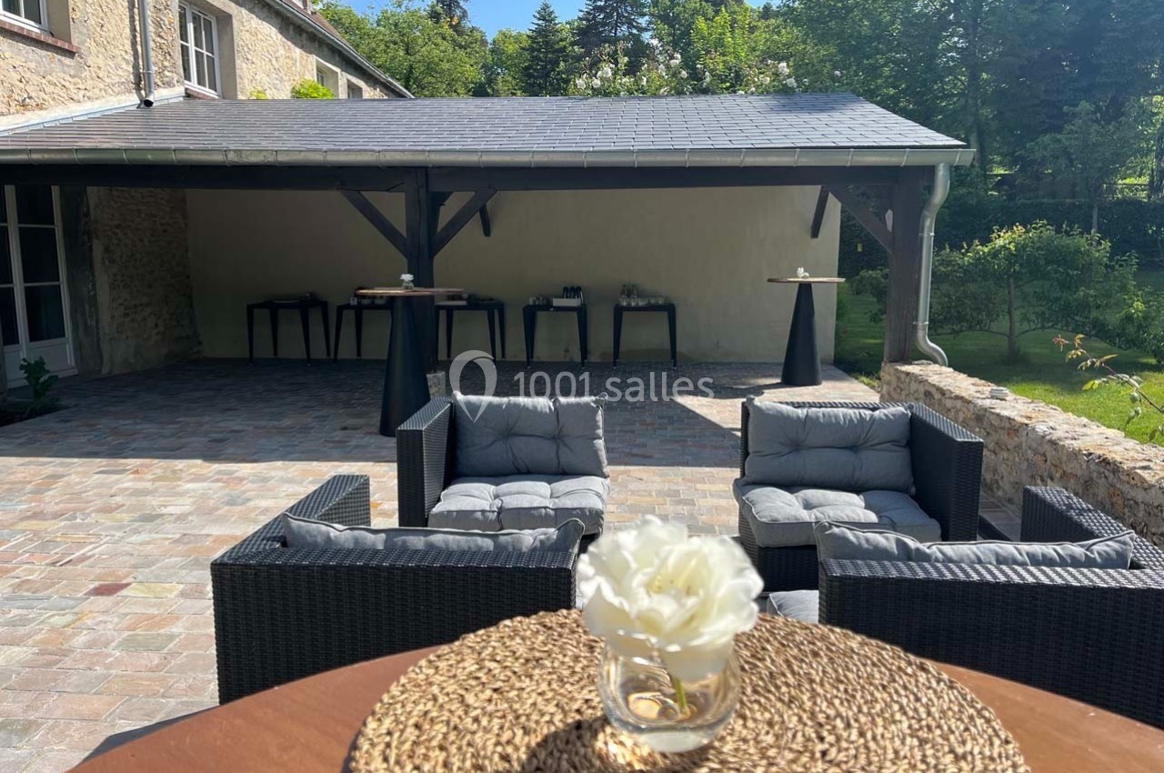 Terrasse en pavés avec fauteuils en rotin, table basse et abri en arrière-plan entouré de verdure.