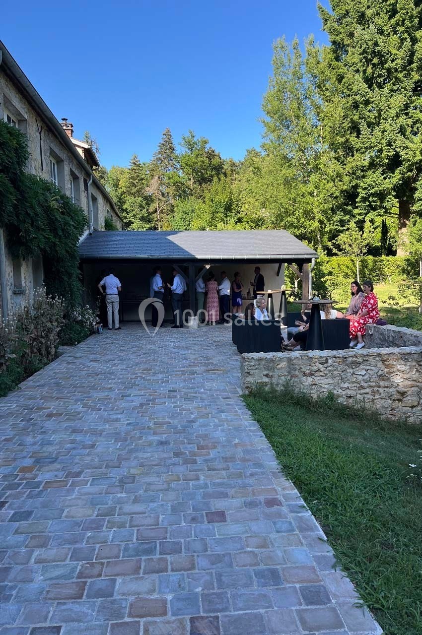 Cour pavée devant une maison en pierre, avec des personnes rassemblées près d'une terrasse couverte et un jardin verdoyant.