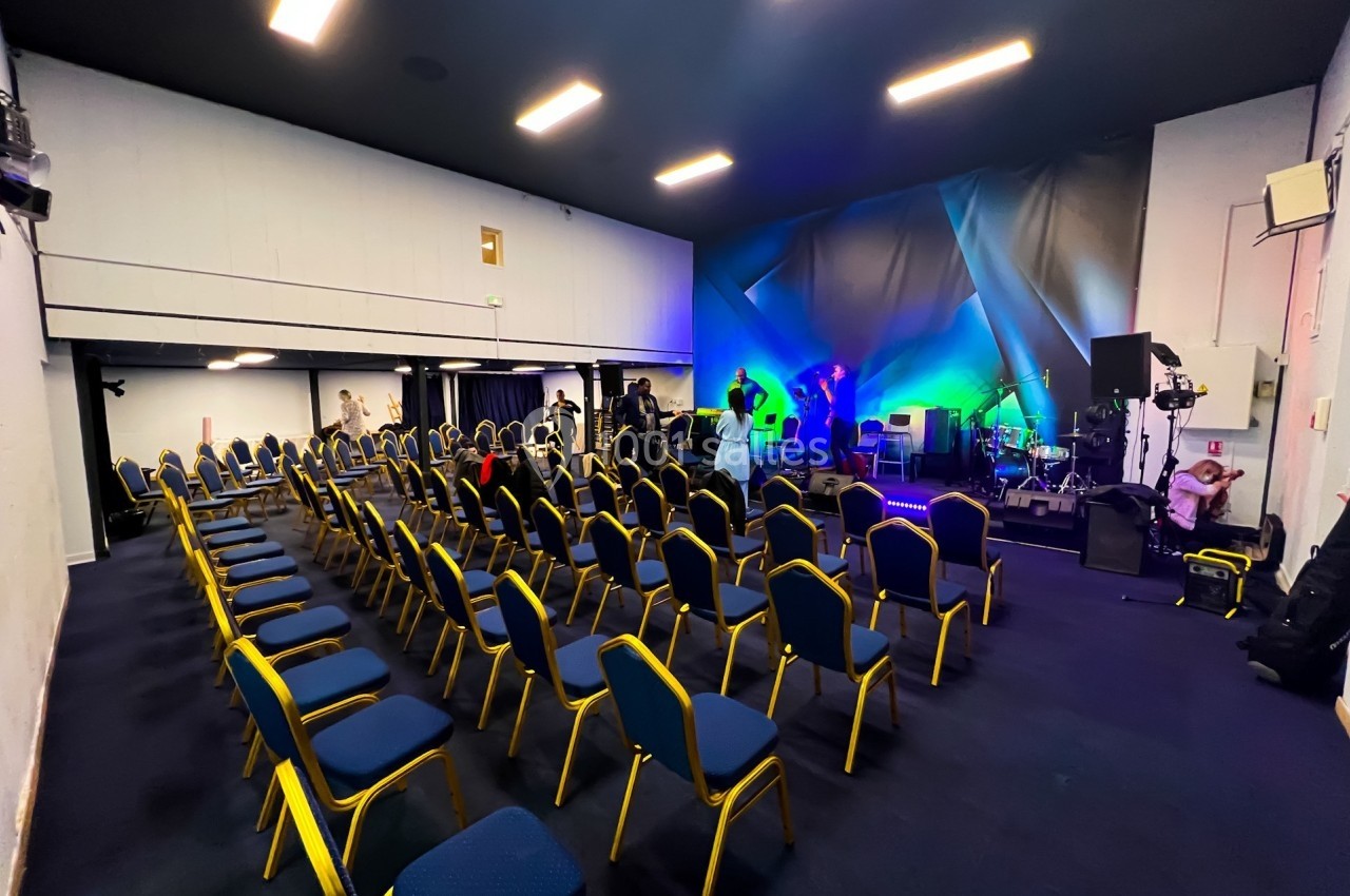 Salle de conférence avec des rangées de chaises vides et une scène équipée de matériel audio et éclairages colorés.