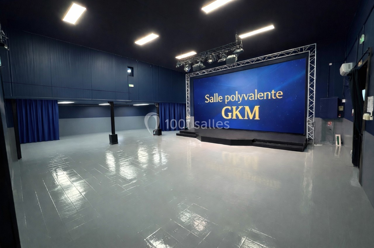 Salle polyvalente avec un grand écran affichant ’Salle polyvalente GKM’, sol brillant et éclairage au plafond.