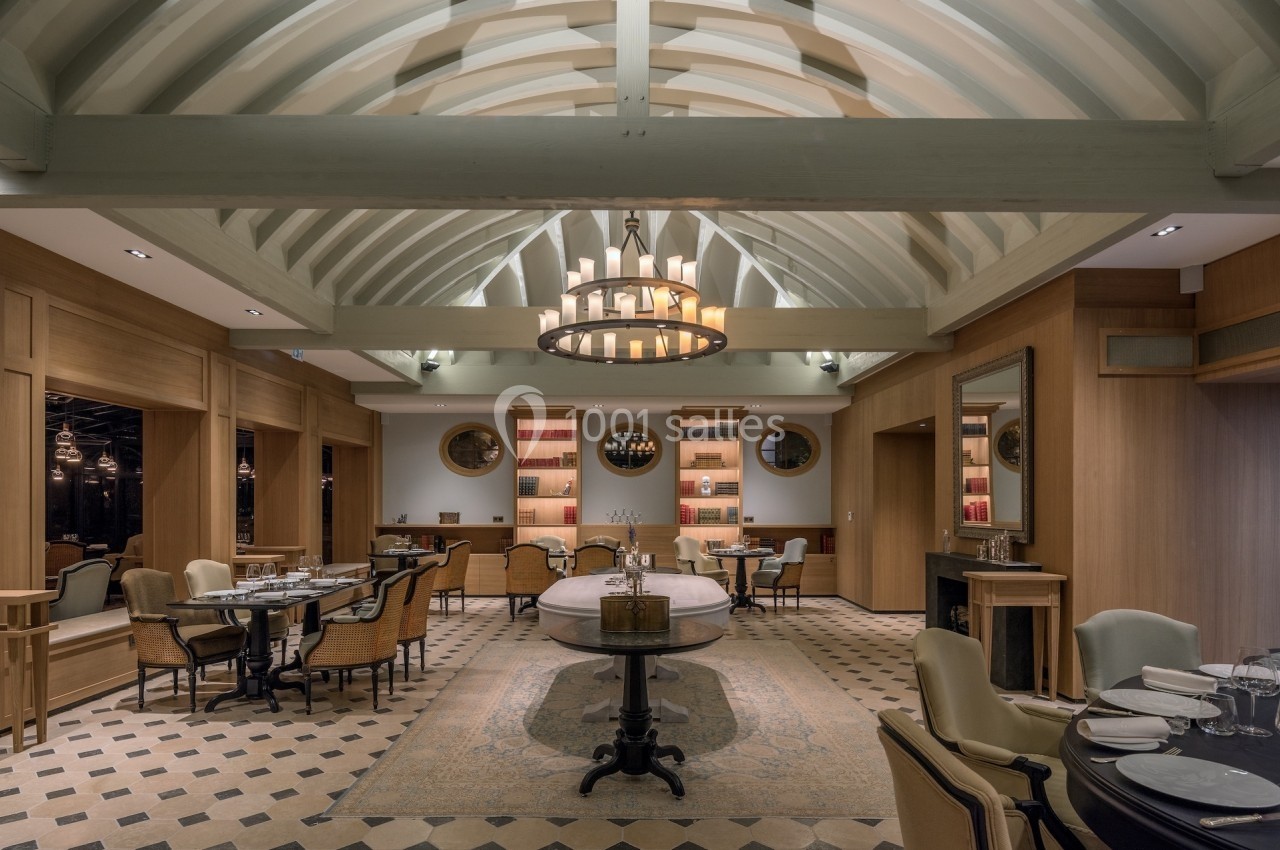 Salle de restaurant élégante avec poutres apparentes, lustre central, tables dressées et ambiance chaleureuse.