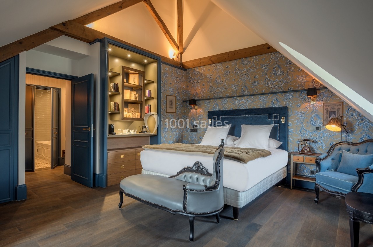 Chambre élégante avec poutres apparentes, lit double, fauteuils, éclairage doux et étagères intégrées.