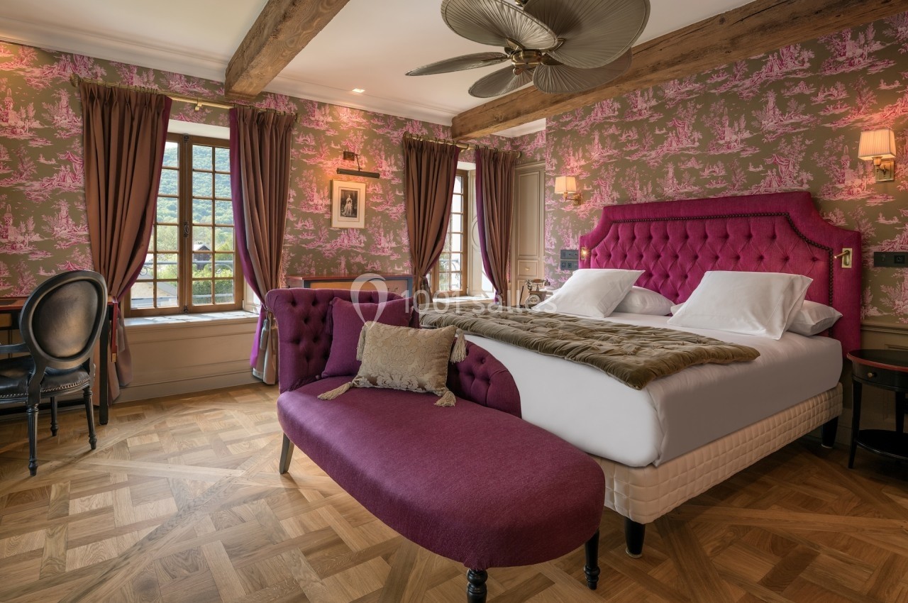 Chambre élégante avec lit double, canapé violet, parquet en bois et fenêtres encadrées de rideaux.
