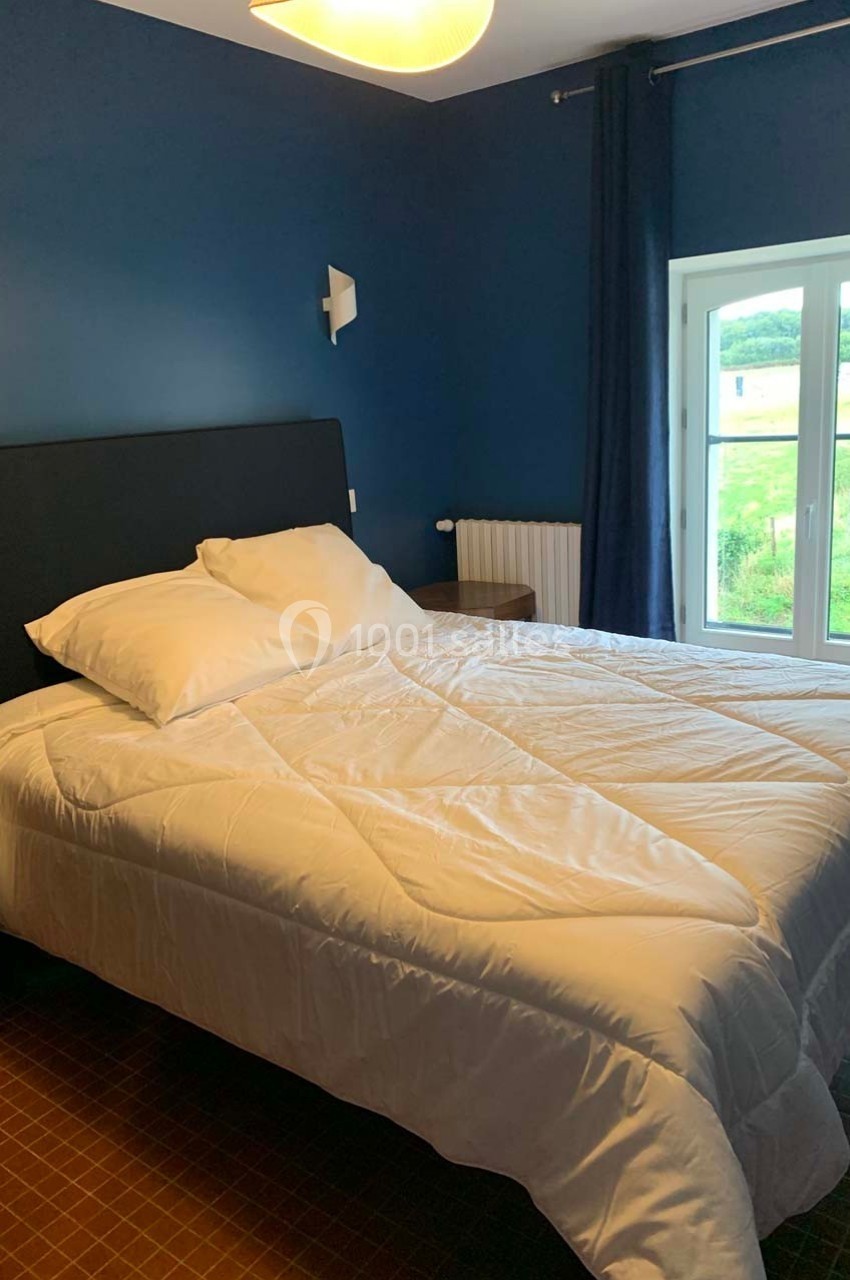 Chambre avec un lit double, murs bleus, fenêtre donnant sur un paysage verdoyant et lumière naturelle.