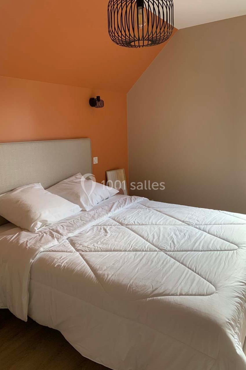 Lit double avec draps blancs dans une chambre aux murs orange et beige, éclairée par une fenêtre et une suspension noire.