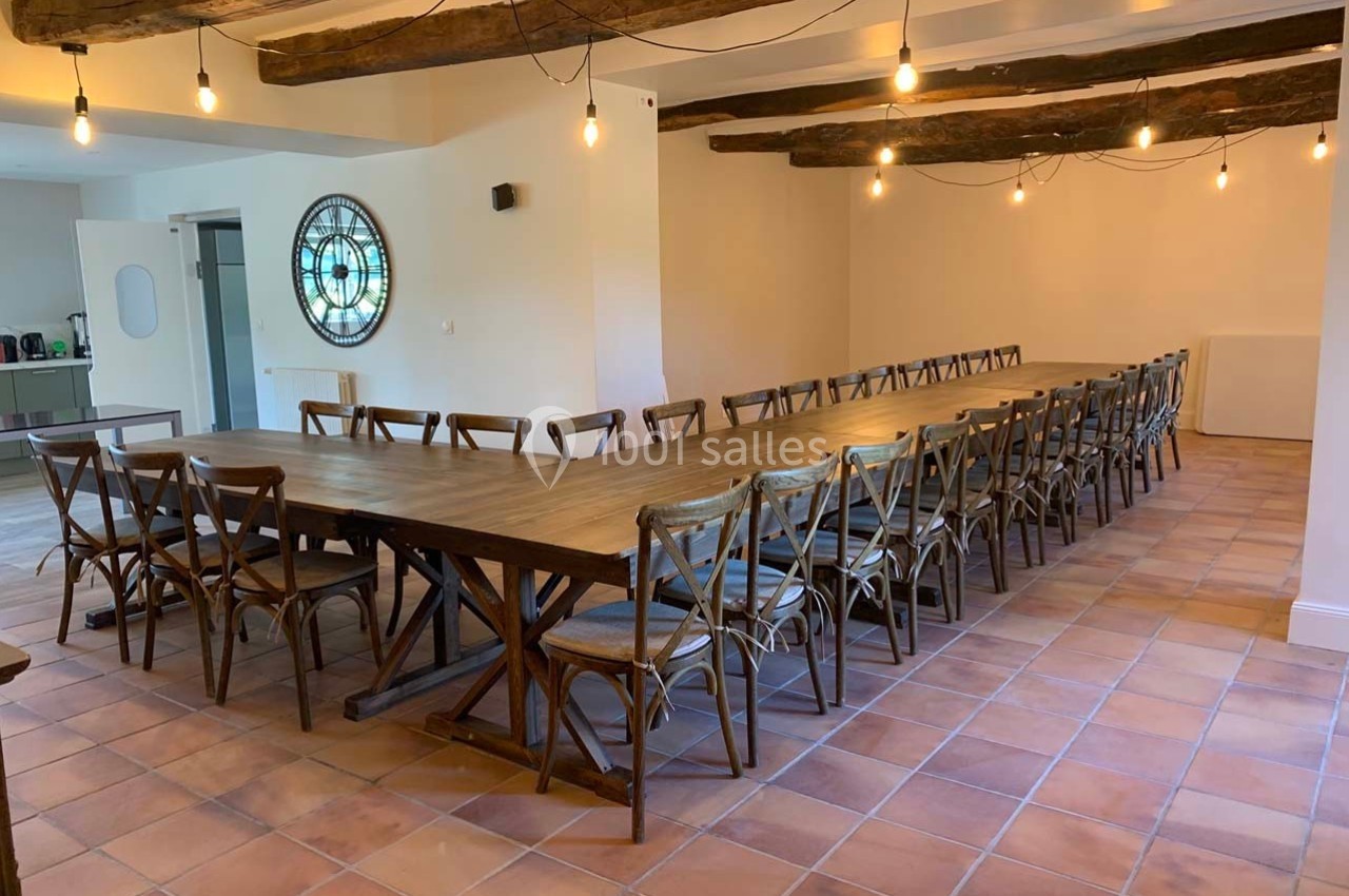 Grande salle avec une longue table en bois entourée de chaises, éclairée par des ampoules suspendues et un vitrail rond.