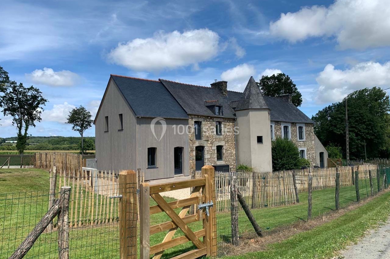 Maison en pierre avec une extension moderne, entourée de clôtures en bois et située dans un paysage rural verdoyant.