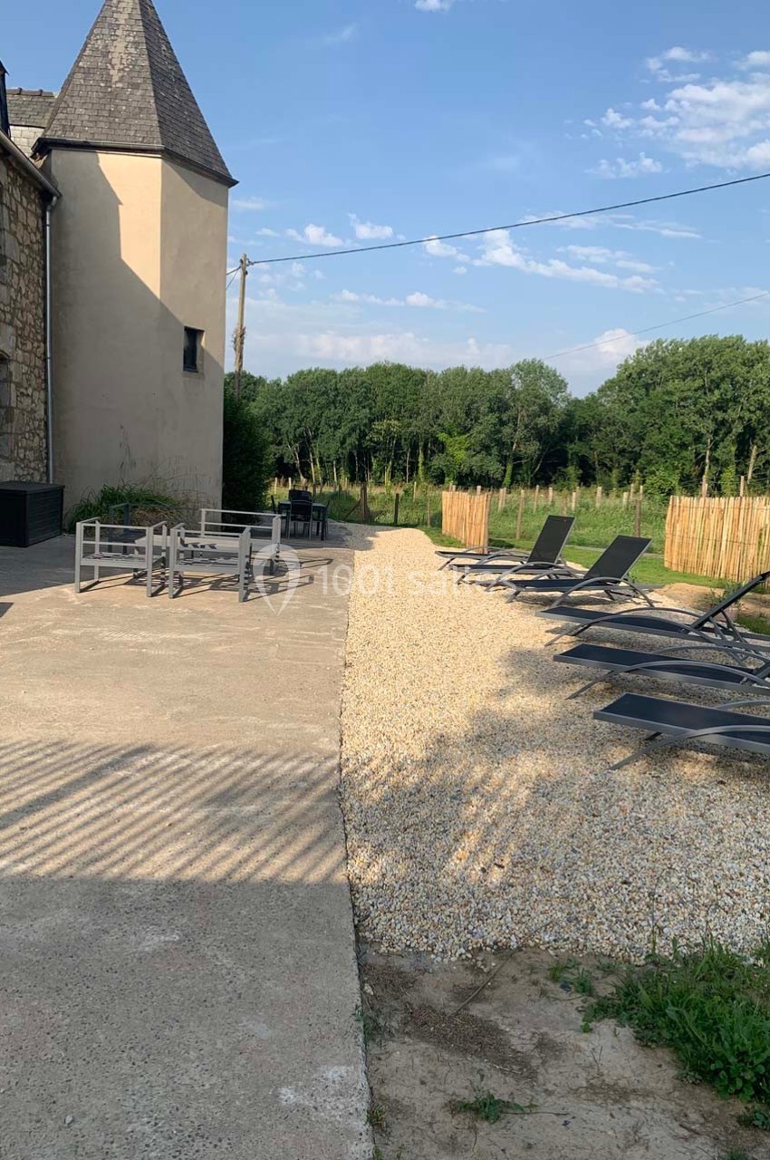 Cour extérieure avec terrasse en gravier, chaises longues, table avec chaises, et vue sur un jardin bordé d'arbres.