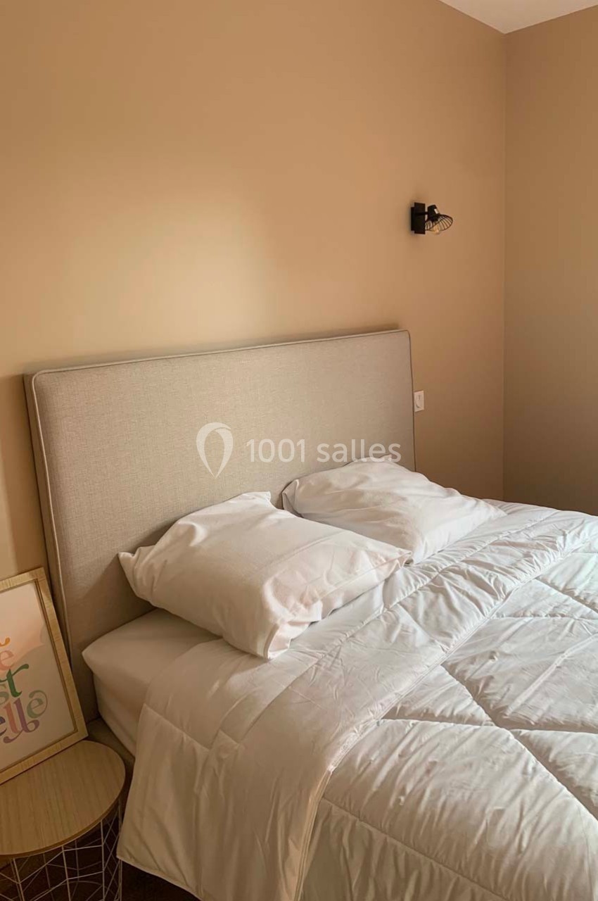 Lit double avec tête de lit beige, draps blancs, table de chevet en bois et cadre décoratif dans une chambre beige.