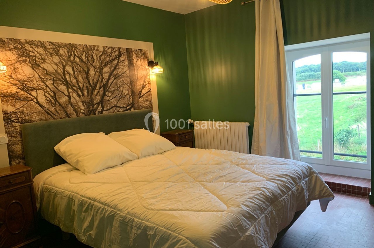 Chambre avec un lit double, murs verts, grande fenêtre donnant sur un paysage champêtre et éclairage doux.