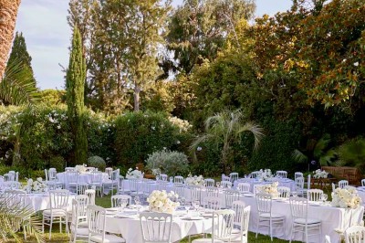 Coussin brodé ’Villa Alexandra’ au premier plan, banc en bois flou en arrière-plan dans un jardin ensoleillé.