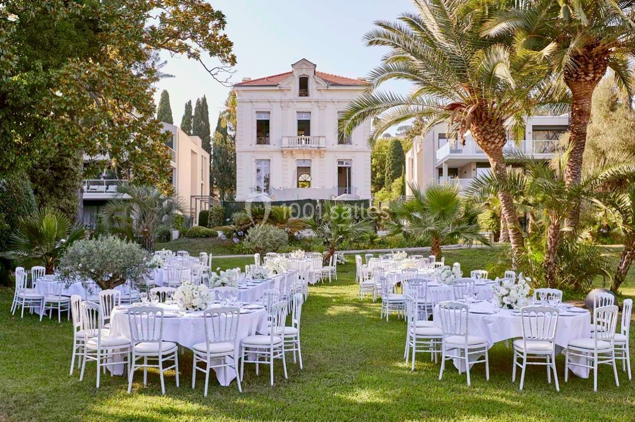 Tables dressées avec nappes blanches et chaises disposées dans un jardin verdoyant devant une grande maison ancienne.