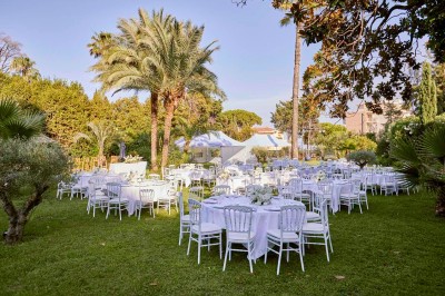 Coussin brodé ’Villa Alexandra’ au premier plan, banc en bois flou en arrière-plan dans un jardin ensoleillé.