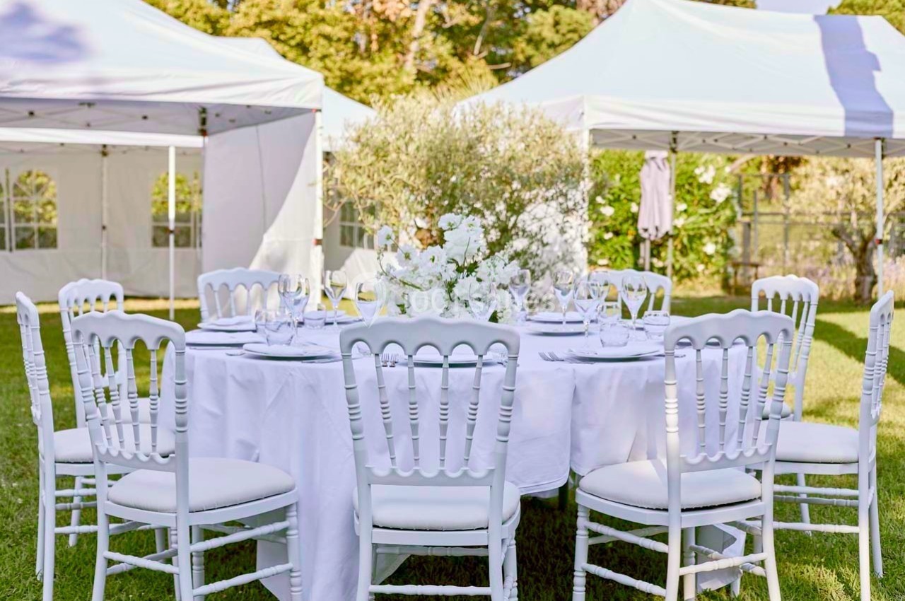 Table ronde dressée avec nappes blanches, chaises assorties et centre de table floral, entourée de tentes blanches en…