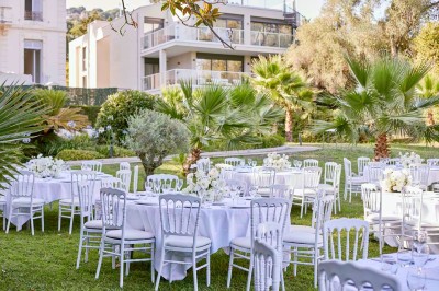 Coussin brodé ’Villa Alexandra’ au premier plan, banc en bois flou en arrière-plan dans un jardin ensoleillé.