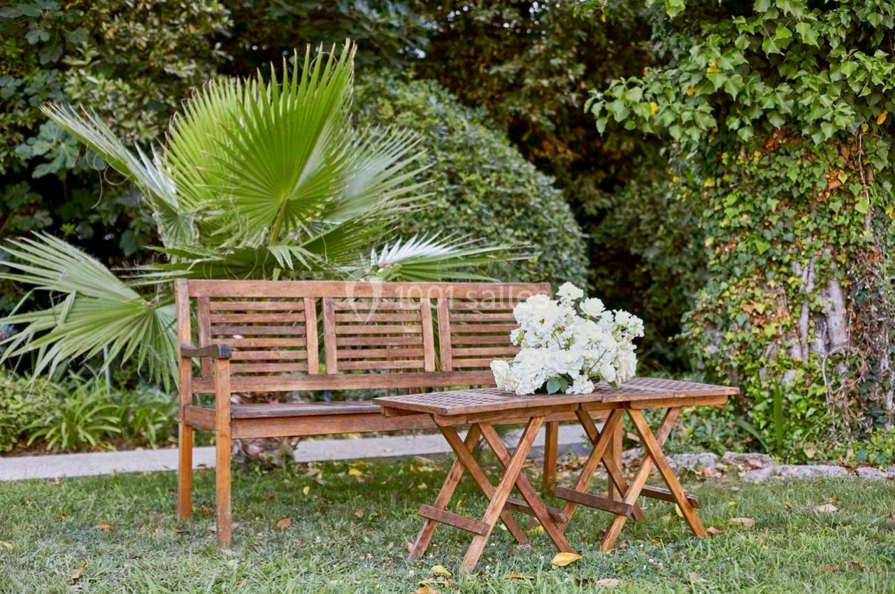 Banc en bois et table assortie avec un bouquet de fleurs blanches, situés dans un jardin verdoyant.