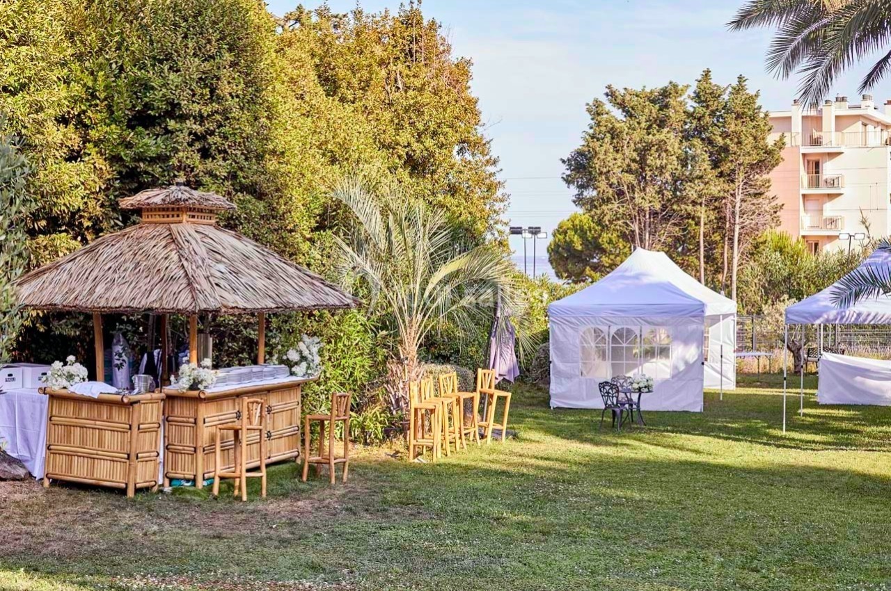 Espace extérieur avec bar en bambou, tentes blanches, tables et chaises, entouré de végétation et d'arbres.