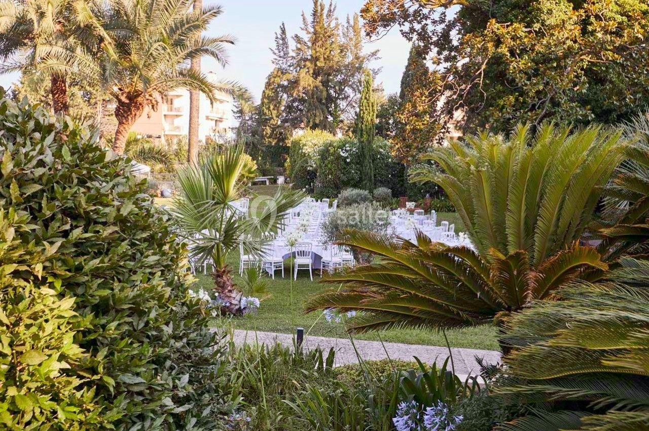 Jardin aménagé avec des tables et chaises blanches disposées pour un événement en plein air, entouré de végétation…