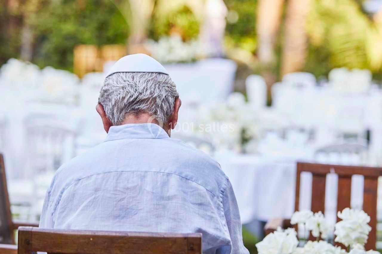 Un homme assis de dos, portant une kippa blanche, dans un jardin avec des tables décorées de fleurs blanches.