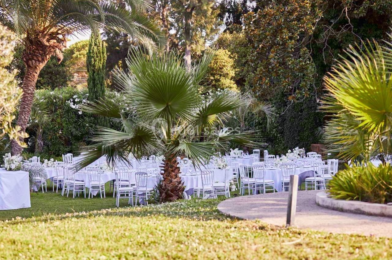 Tables et chaises blanches disposées en extérieur dans un jardin verdoyant, entourées de palmiers et de végétation.