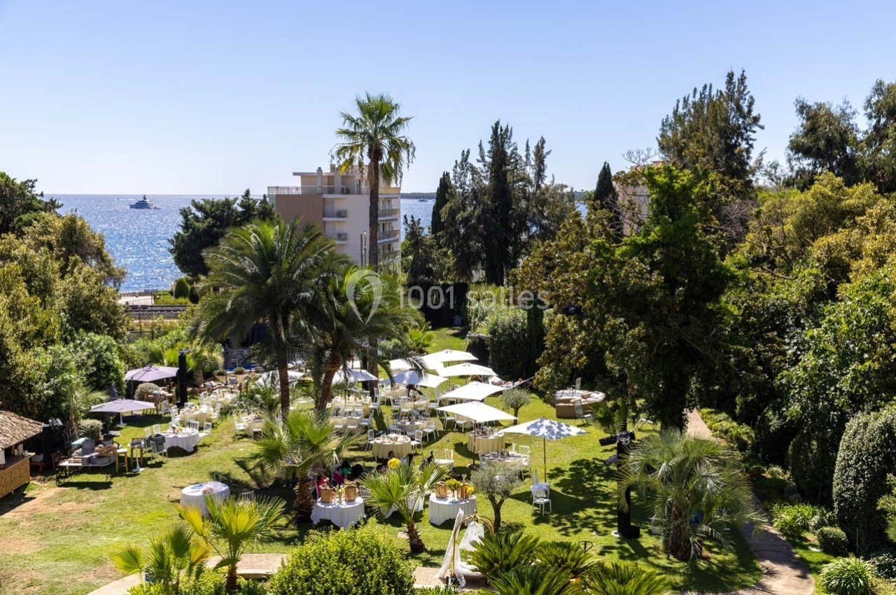 Jardin avec tables dressées pour un repas en plein air, entouré de palmiers, avec vue sur la mer et un bâtiment en arrière…