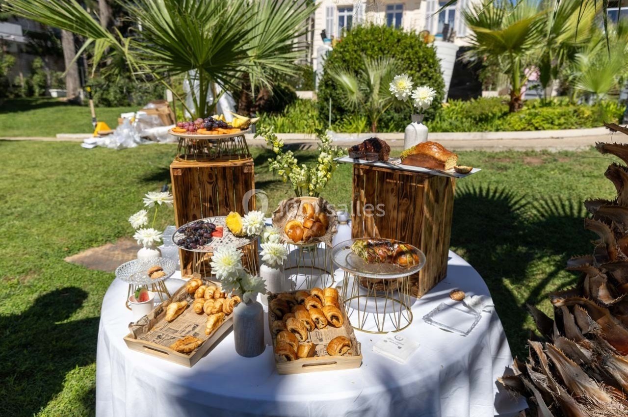 Table ronde en extérieur avec des pâtisseries variées présentées sur des supports en bois, entourée de verdure.