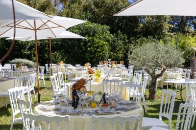 Coussin brodé ’Villa Alexandra’ au premier plan, banc en bois flou en arrière-plan dans un jardin ensoleillé.