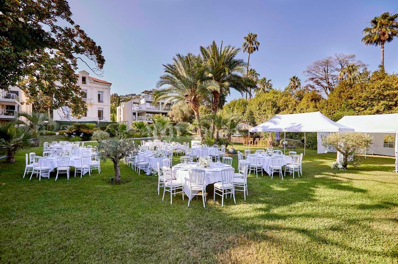 Tables rondes dressées avec des nappes blanches et chaises disposées dans un jardin verdoyant avec des arbres et une tente…