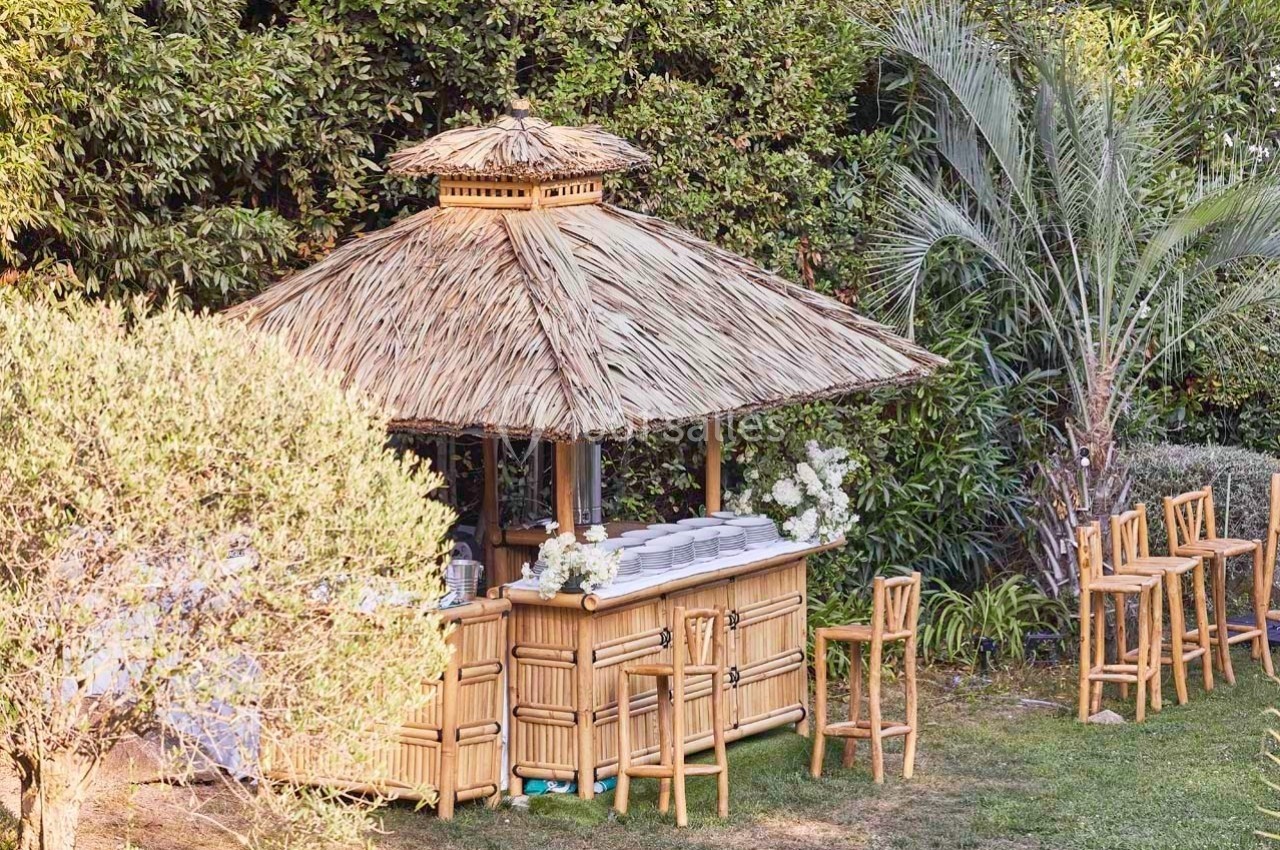 Bar en bambou avec toit en chaume, entouré de chaises en bois et de végétation tropicale dans un jardin.