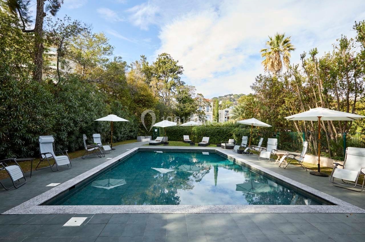Piscine extérieure entourée de chaises longues, parasols blancs et végétation luxuriante sous un ciel partiellement dégagé.