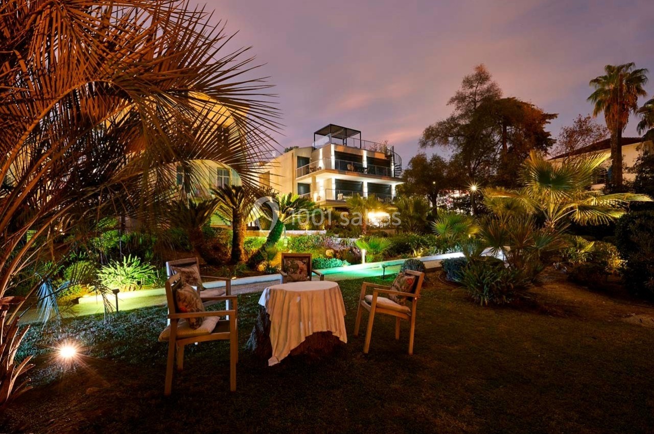 Jardin éclairé au crépuscule avec une table et des chaises au premier plan, et une maison moderne en arrière-plan.