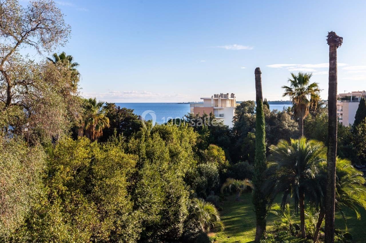 Vue sur un jardin arboré avec des palmiers, des arbres variés et en arrière-plan la mer et des bâtiments clairs.