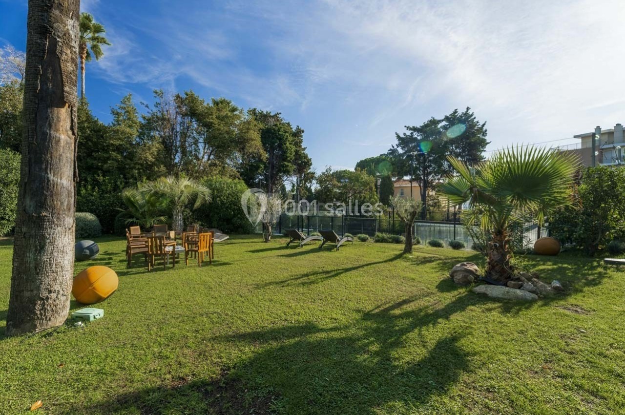 Jardin avec pelouse, table en bois entourée de chaises, arbres et arbustes, sous un ciel bleu dégagé.