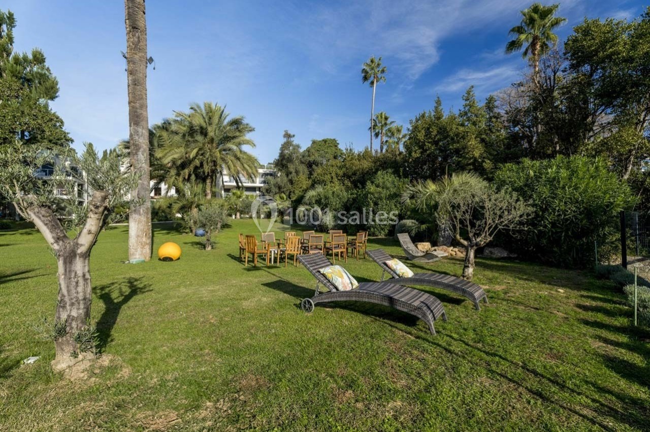 Chaises et tables en bois disposées sur une pelouse entourée d'arbres, avec des transats au premier plan.