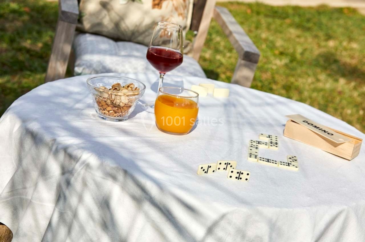 Table ronde en extérieur avec un verre de jus, un verre de vin, des noix, un jeu de dominos et une nappe blanche.