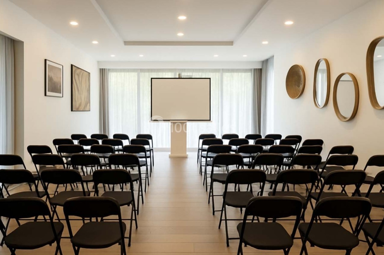 Salle de conférence lumineuse avec des rangées de chaises noires face à un écran blanc sur un support.