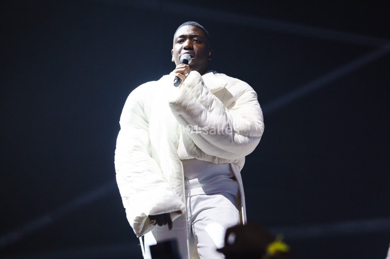 Un chanteur en manteau blanc se produit sur scène, tenant un micro sous un éclairage tamisé.