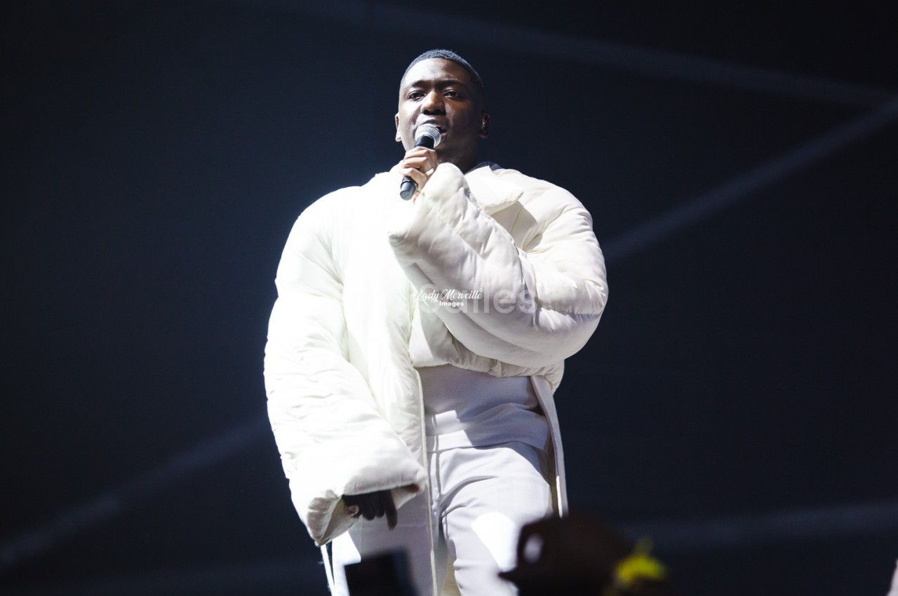 Un chanteur en manteau blanc se produit sur scène, tenant un micro sous un éclairage tamisé.