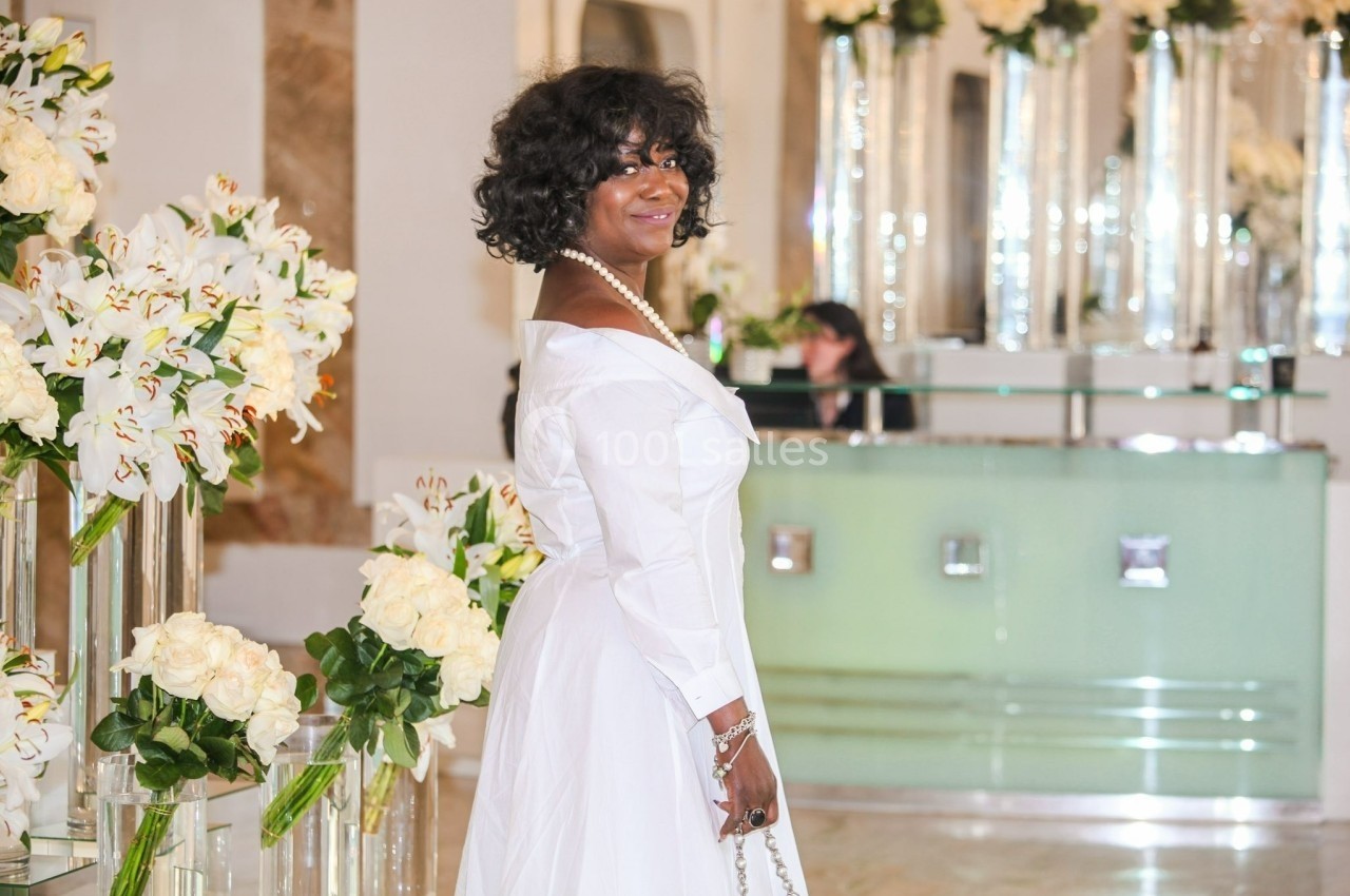 Femme en robe blanche posant dans un intérieur élégant décoré de fleurs blanches et de vases en verre.