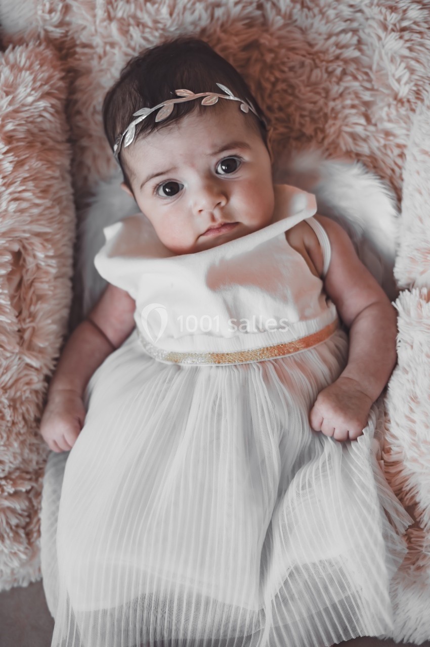 Bébé allongé sur une couverture douce, vêtu d'une robe blanche et d'un bandeau décoratif.