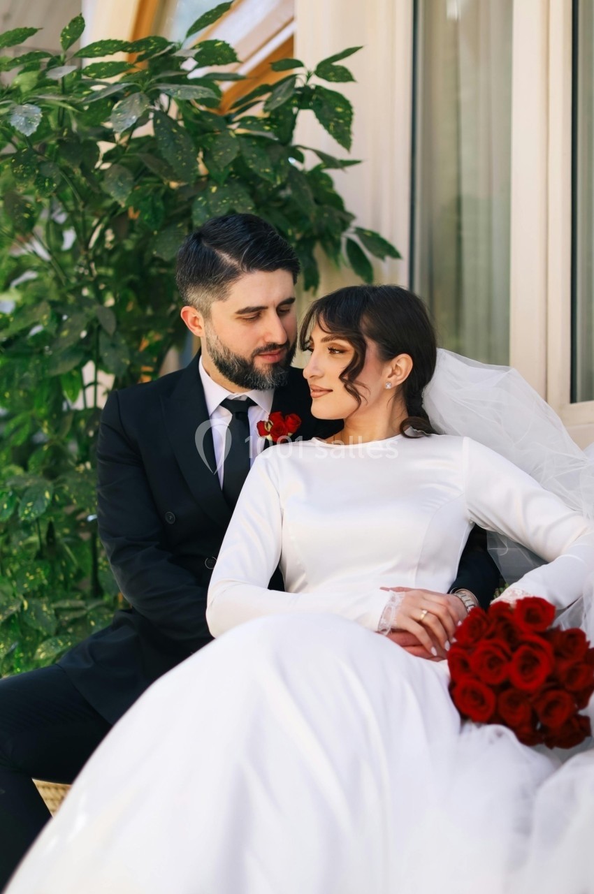 Un couple de mariés assis, la mariée en robe blanche tenant un bouquet de roses rouges, près d'une plante verte.