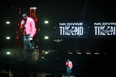 Un chanteur sur scène, vêtu de blanc, levant un bras sous des lumières de spectacle et un écran en arrière-plan.